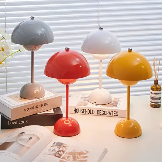 Four colorful table lamps on a surface with books and a bottle in the background.