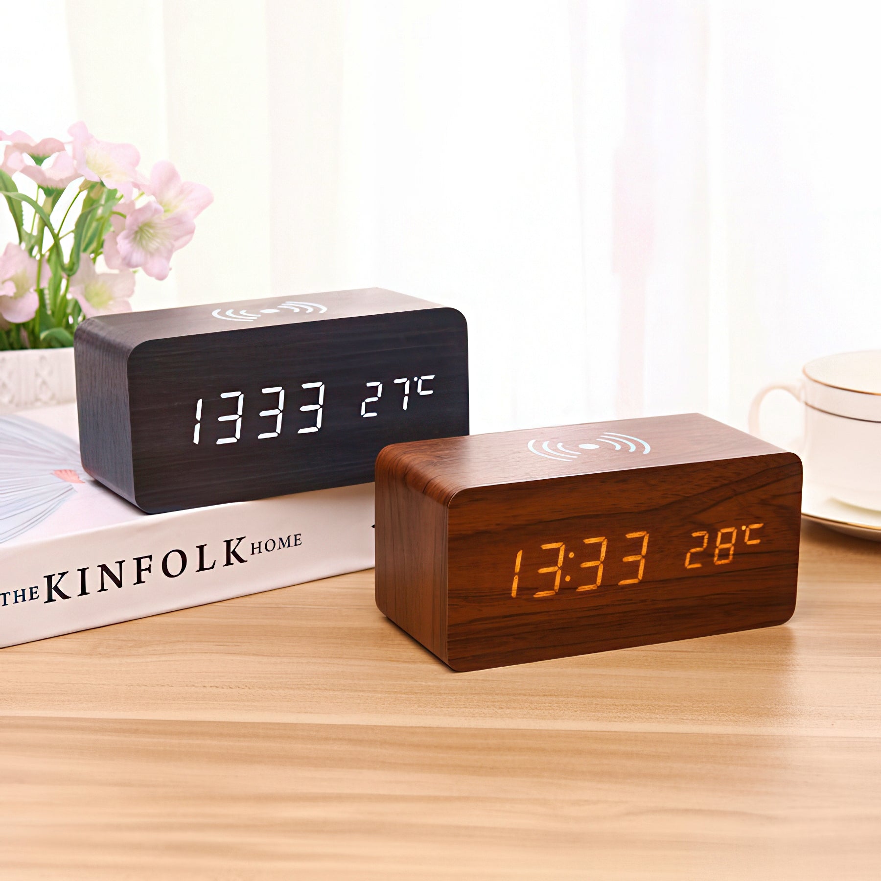 Two digital clocks on a wooden surface with a book and flowers in the background.