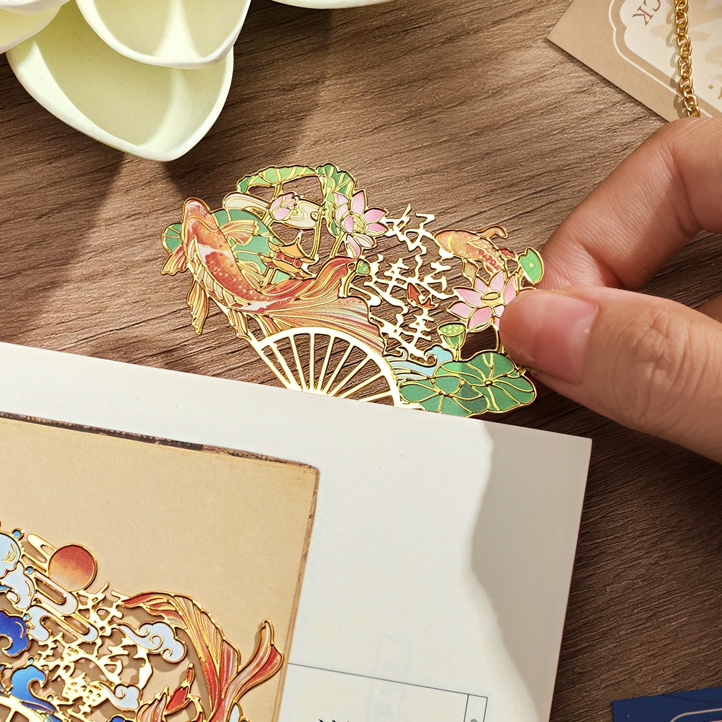 Decorative metal bookmark with floral and fish design held by a hand.