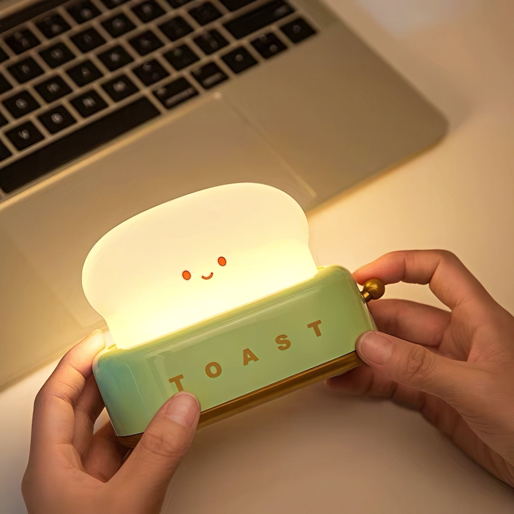 Person holding a cute toaster-shaped night light with a smiling face, illuminated, in front of a laptop.