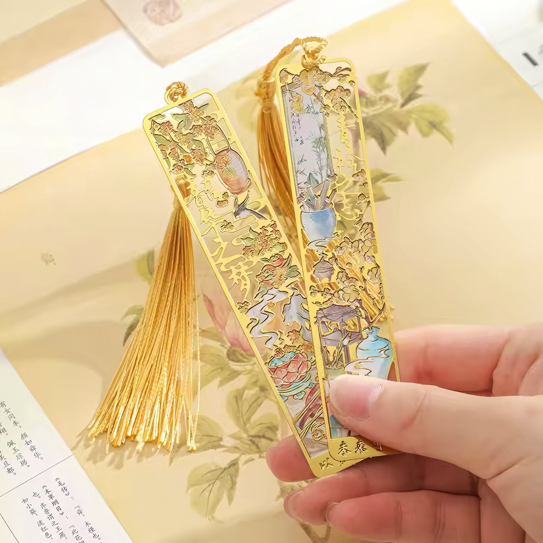 Gold bookmarks with intricate designs held by a hand on a floral-patterned surface.