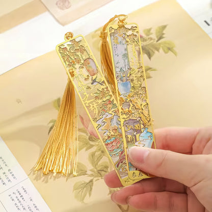 Gold bookmarks with intricate designs held by a hand on a floral-patterned surface.